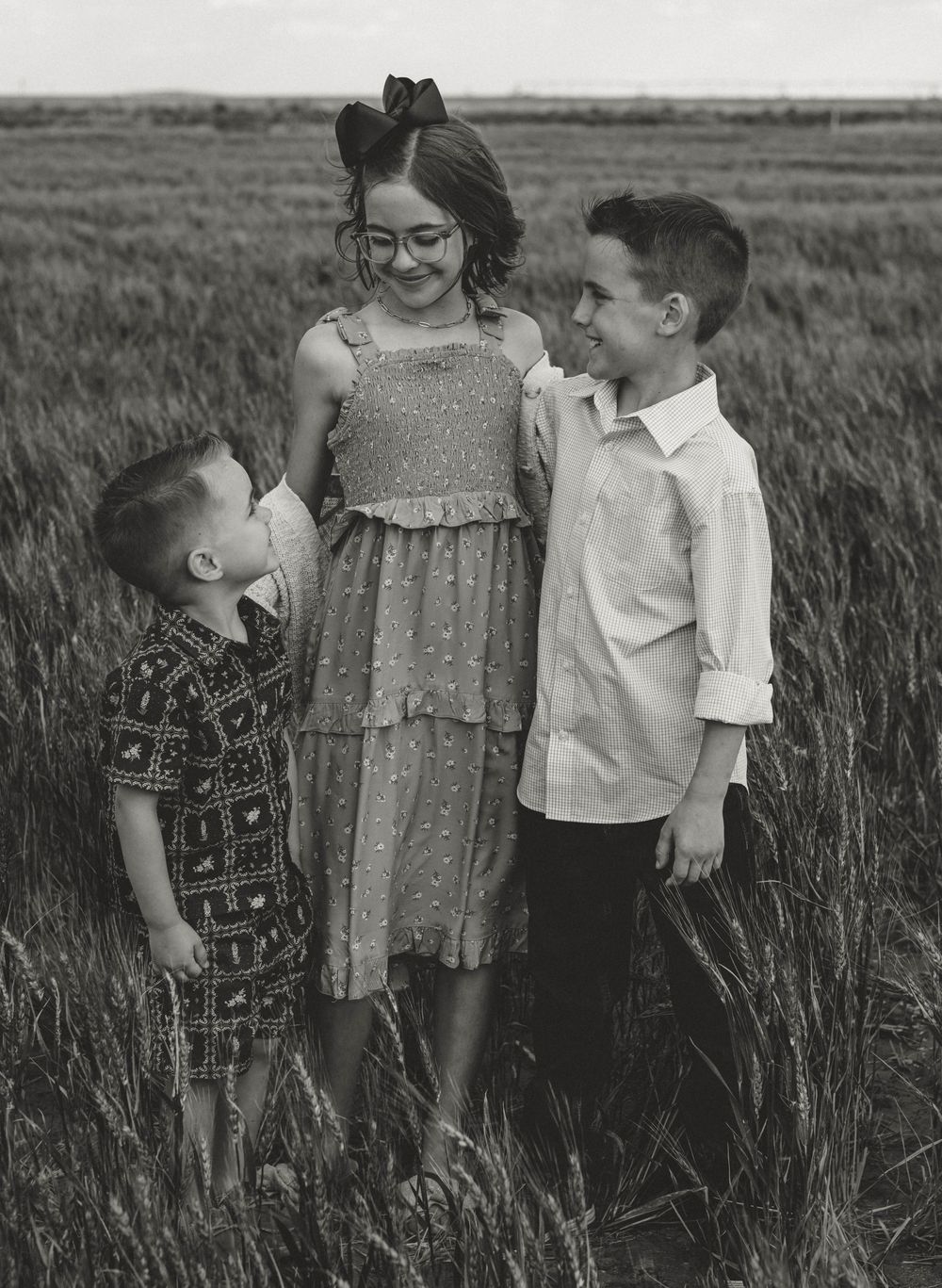 Three children pose together in a wheat field in a black and white photo, with the girl in the middle wearing glasses.