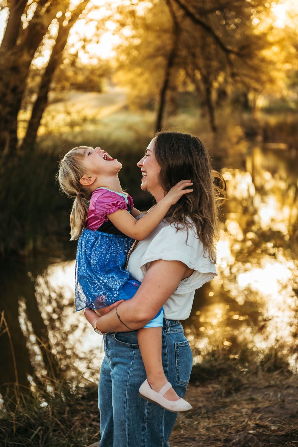 A mother laughs while holding her young daughter outdoors near a sunlit pond during golden hour.
