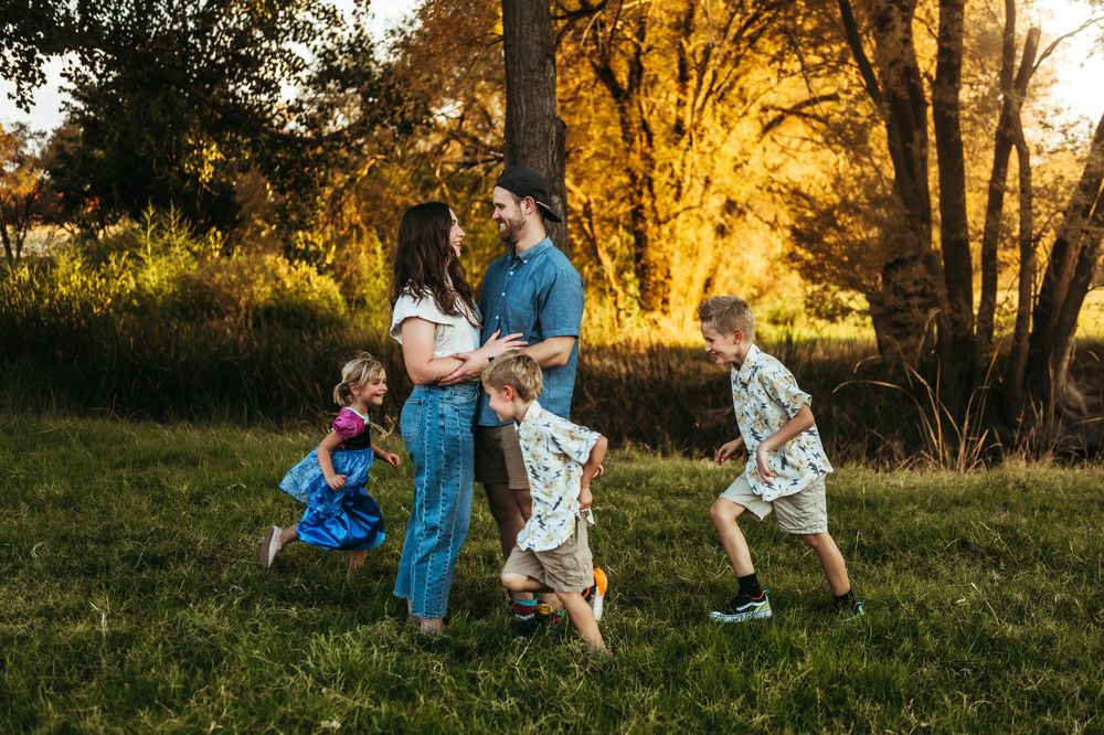Family with three young children playing outdoors in golden sunset light among lush green grass and autumn trees.