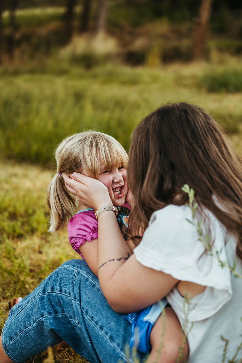 Mother tenderly holding young blonde daughters face while sitting together laughing in grassy field.