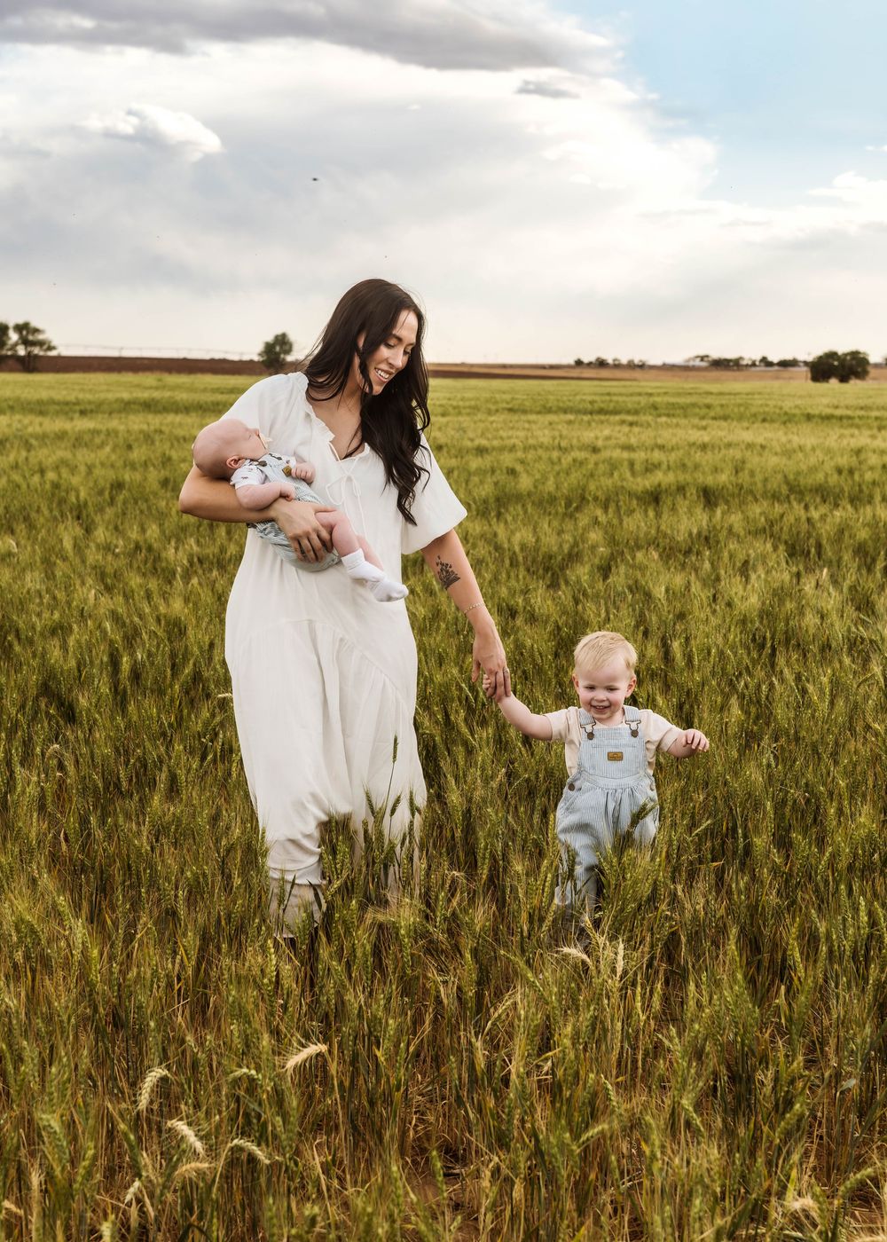 A mother in a white jumpsuit holds a newborn while walking with her toddler in overalls through a green field.