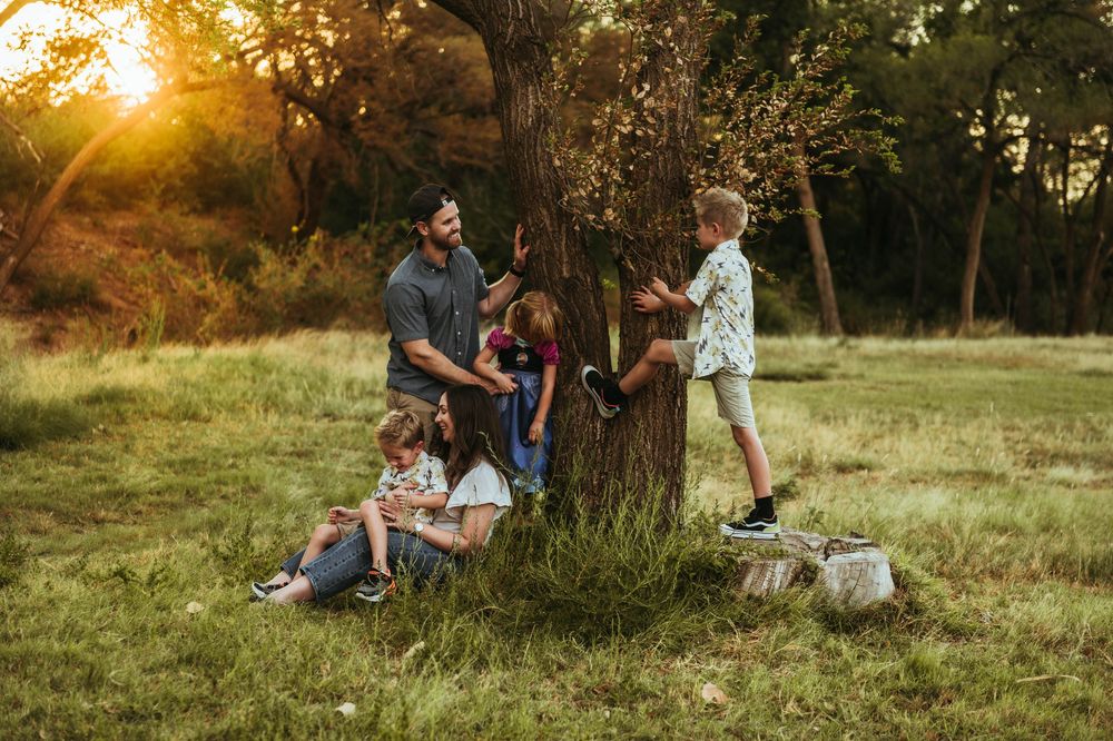 Family gathering around a large tree in a sunlit meadow at golden hour, with children playing and parents watching.