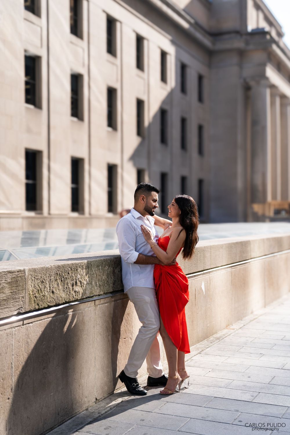 Intimate moment captured against classic stone building with woman in flowing red dress.