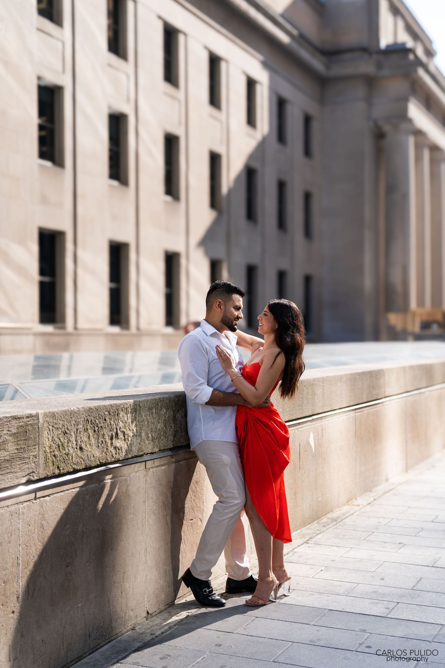 Intimate moment captured against classic stone building with woman in flowing red dress.