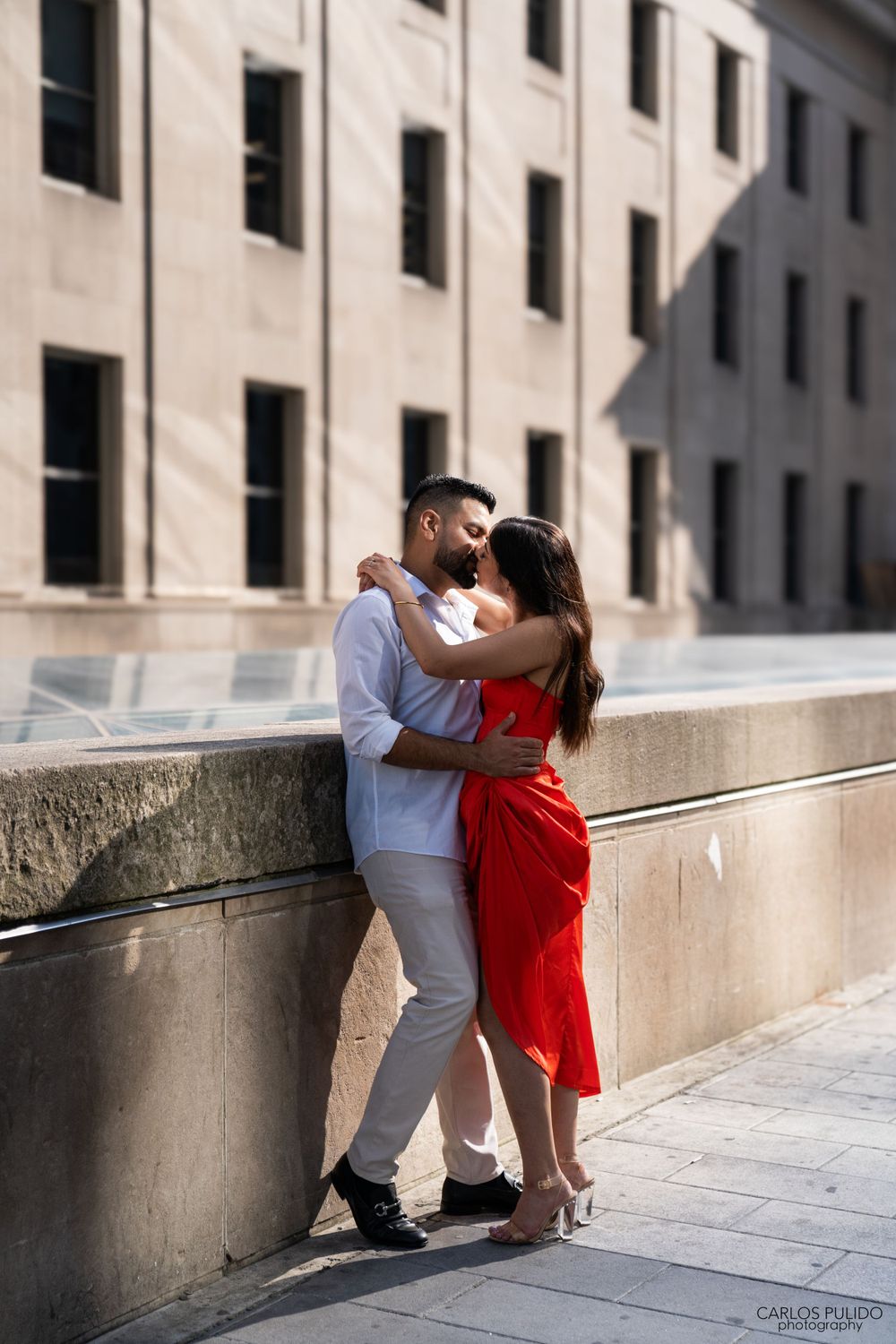 Elegant couple in red dress and white outfit embracing against stone building facade.
