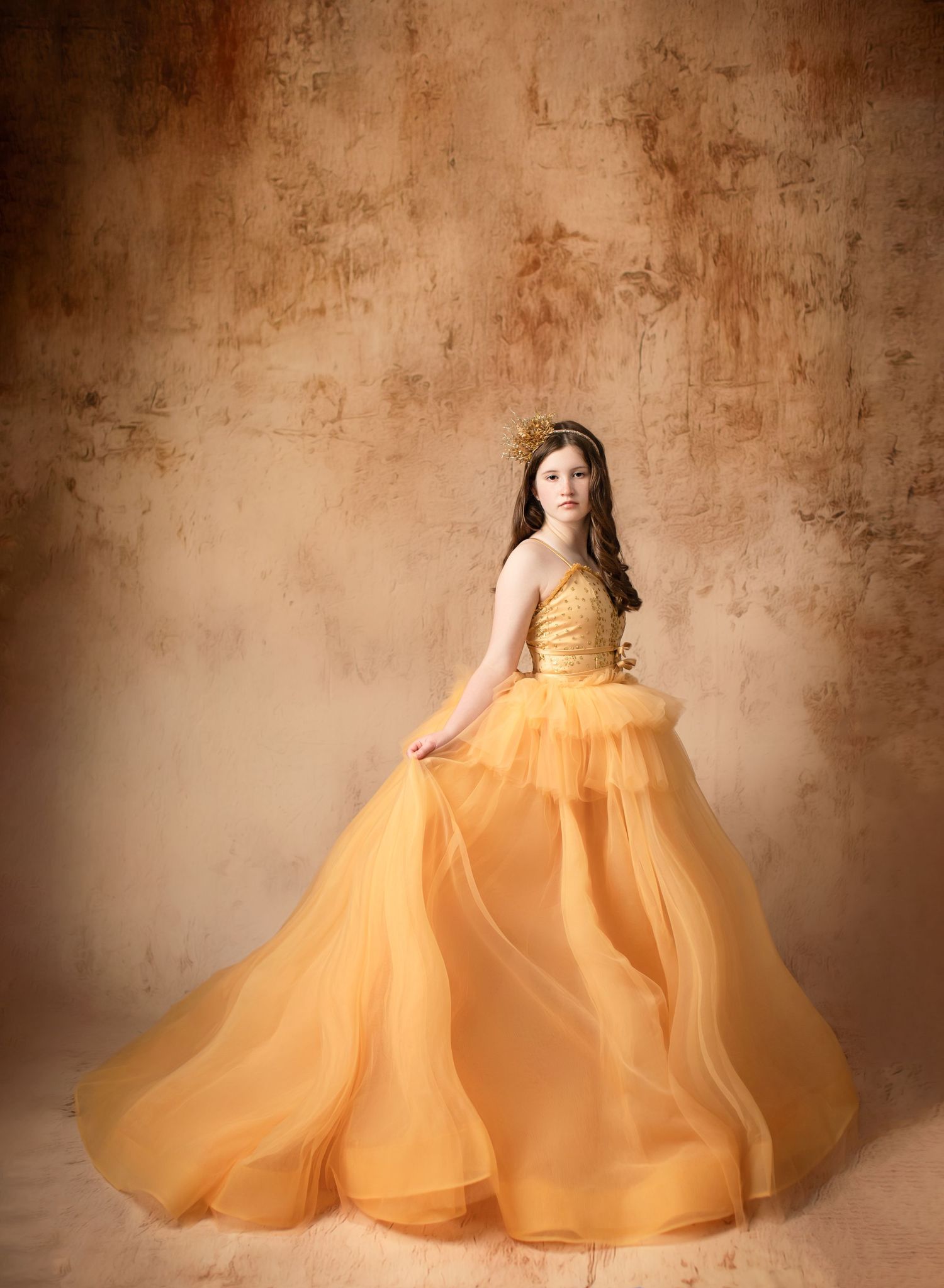 Stunning golden ball gown with layered tulle skirt against textured brown backdrop.