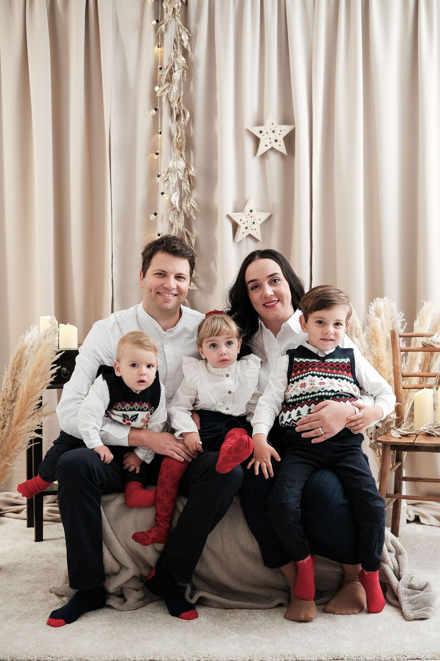 Family group portrait with children wearing coordinated holiday sweaters against cream curtain backdrop.