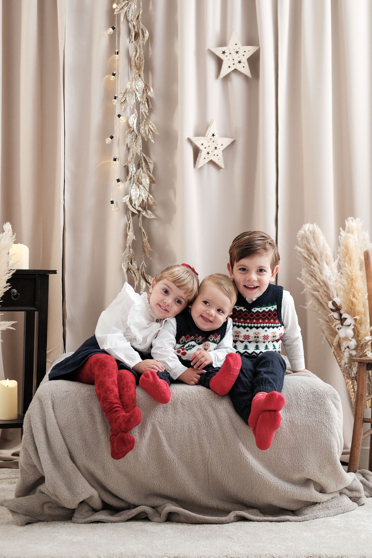 Children lying on grey blanket wearing festive outfits with holiday decorations in background.