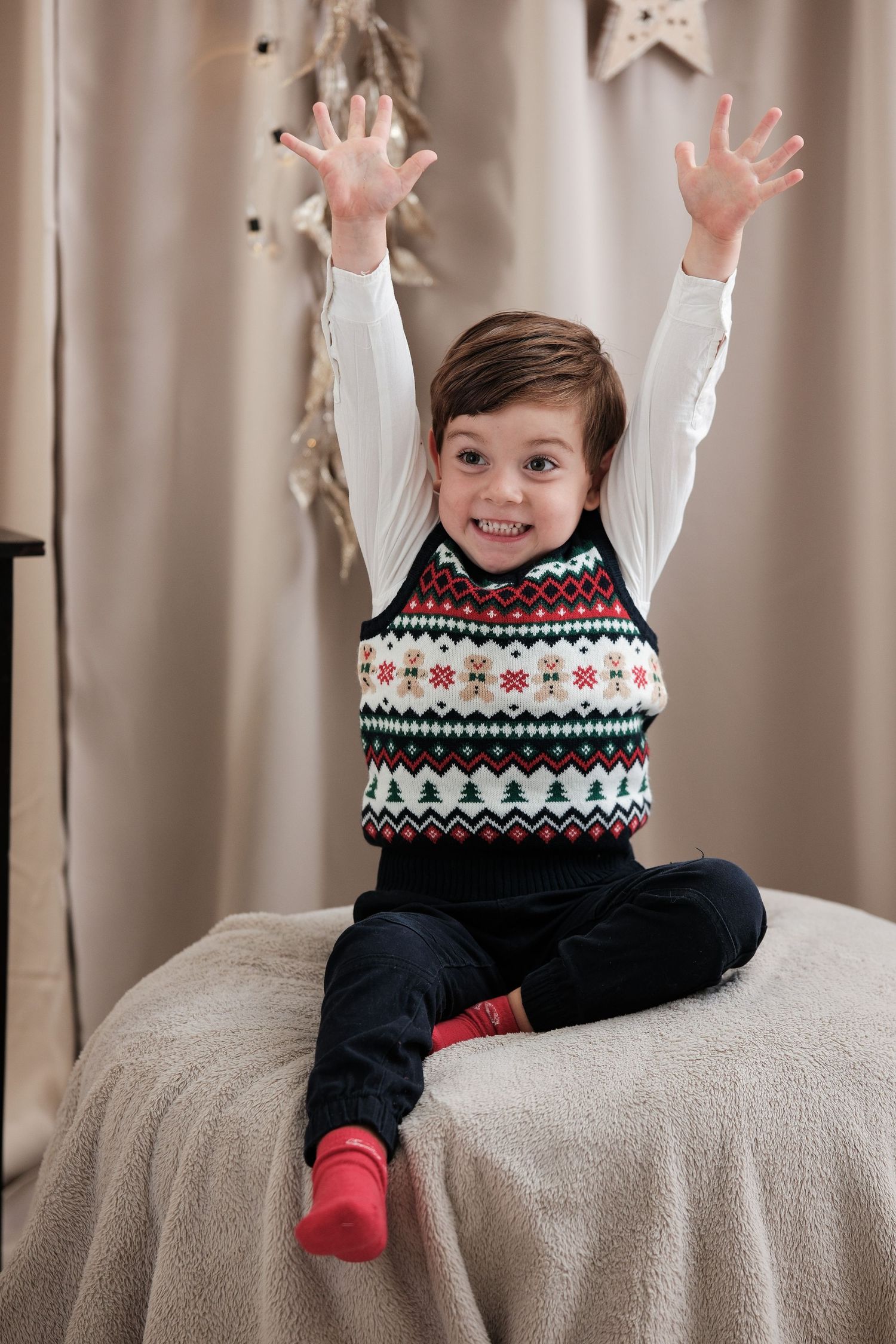 Child in festive fair isle sweater raising arms joyfully on gray couch.