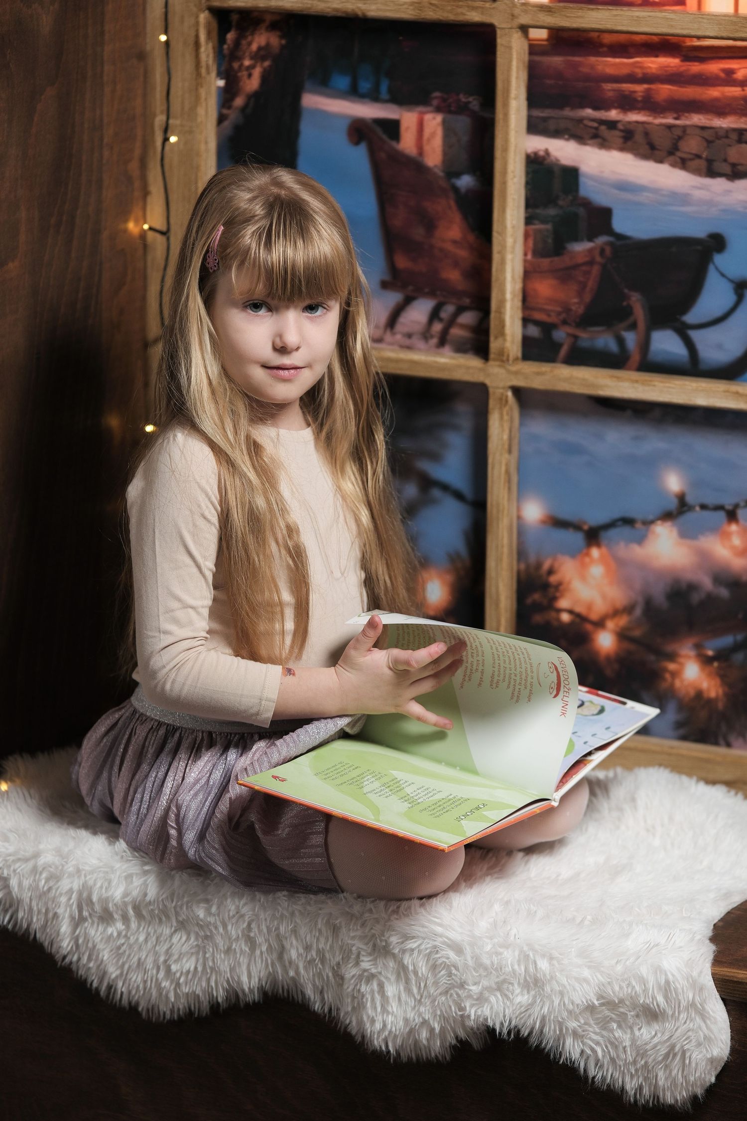 Child reading book on white fur rug by window with winter scene and string lights.