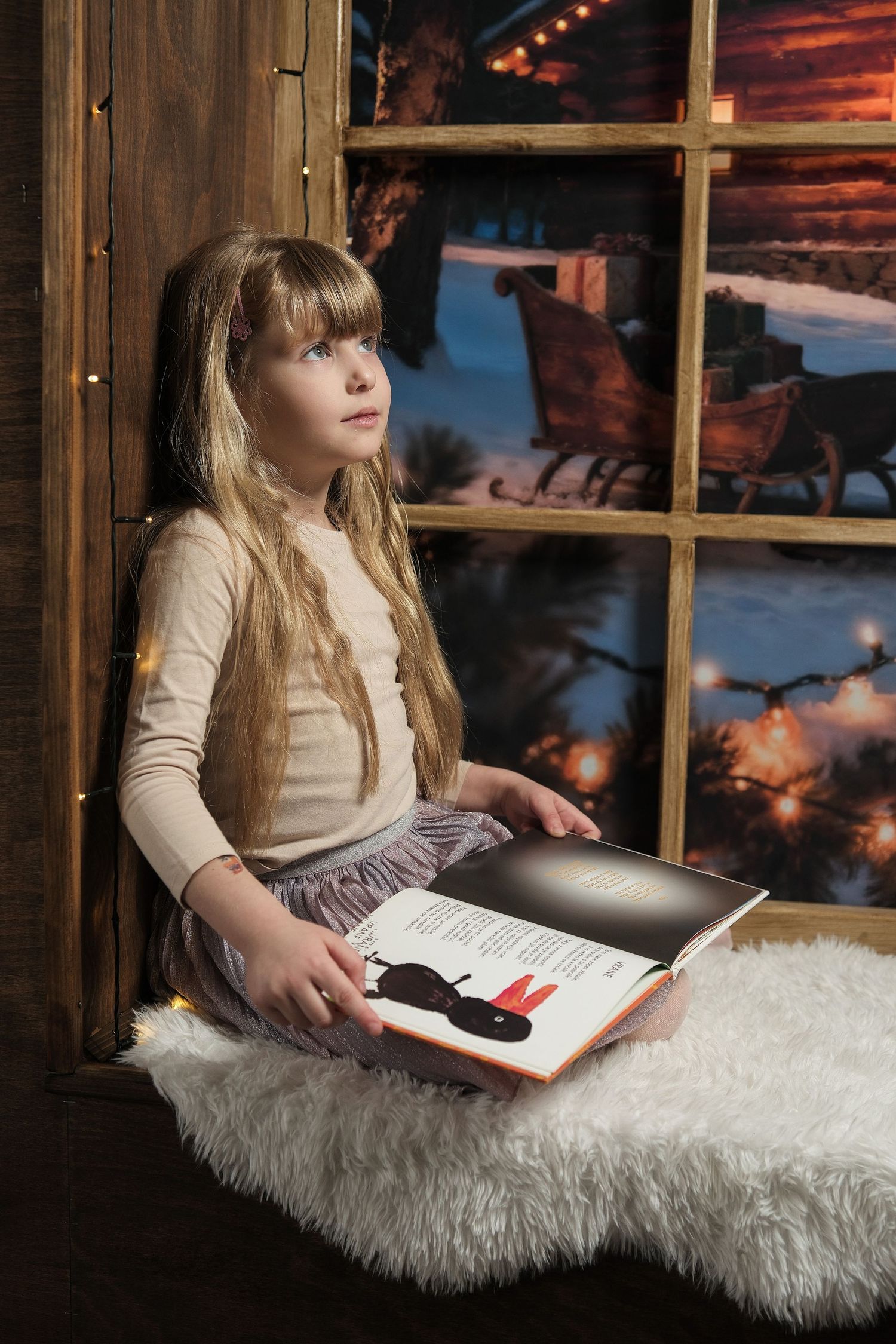 Child looking at book while sitting on fur rug near window with snowy evening scene.