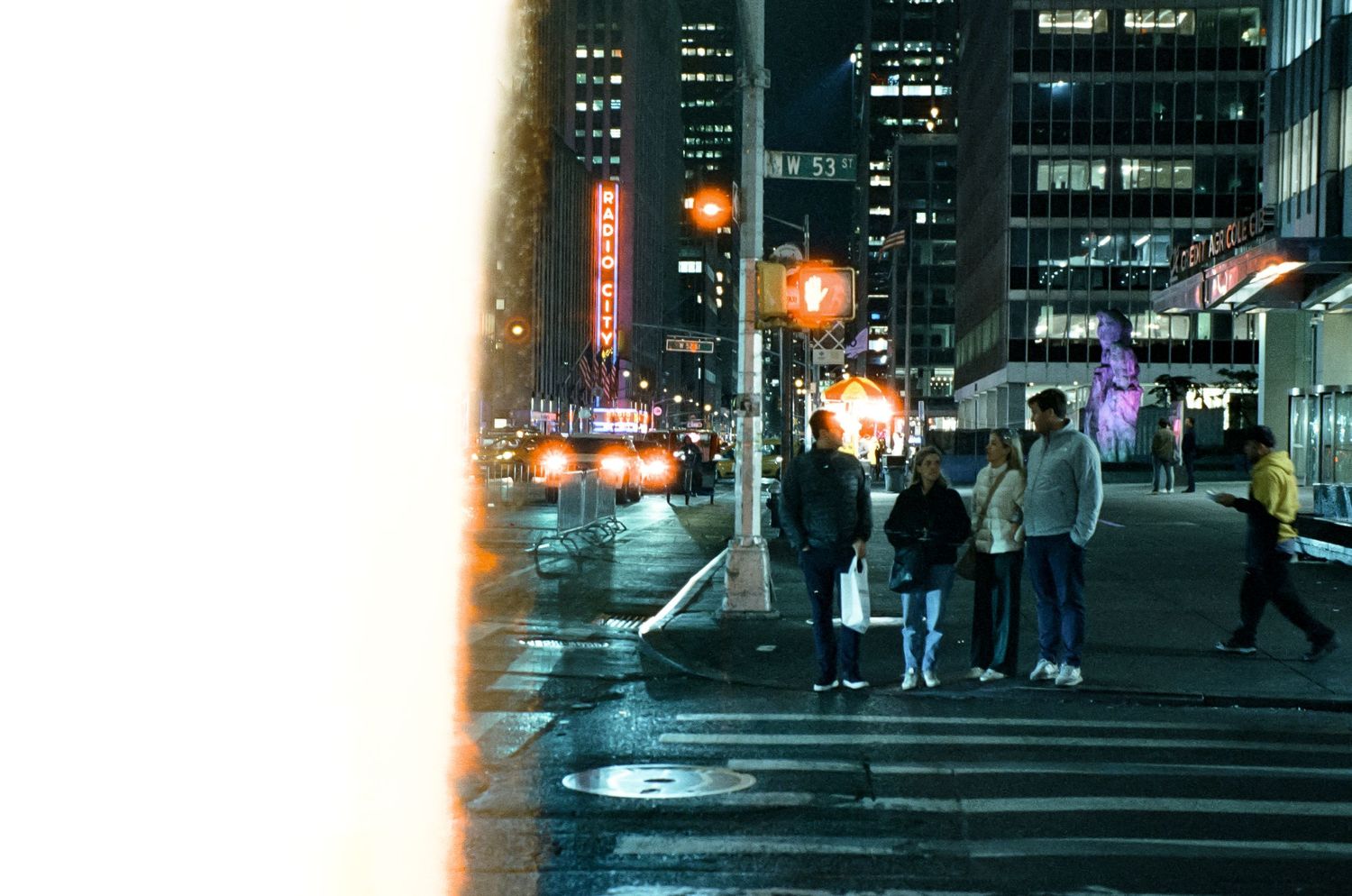 People waiting at a city street corner at night with illuminated buildings in the background.