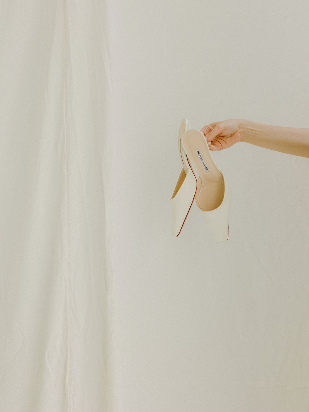A hand holds an elegant cream-colored high heel shoe against a light background.