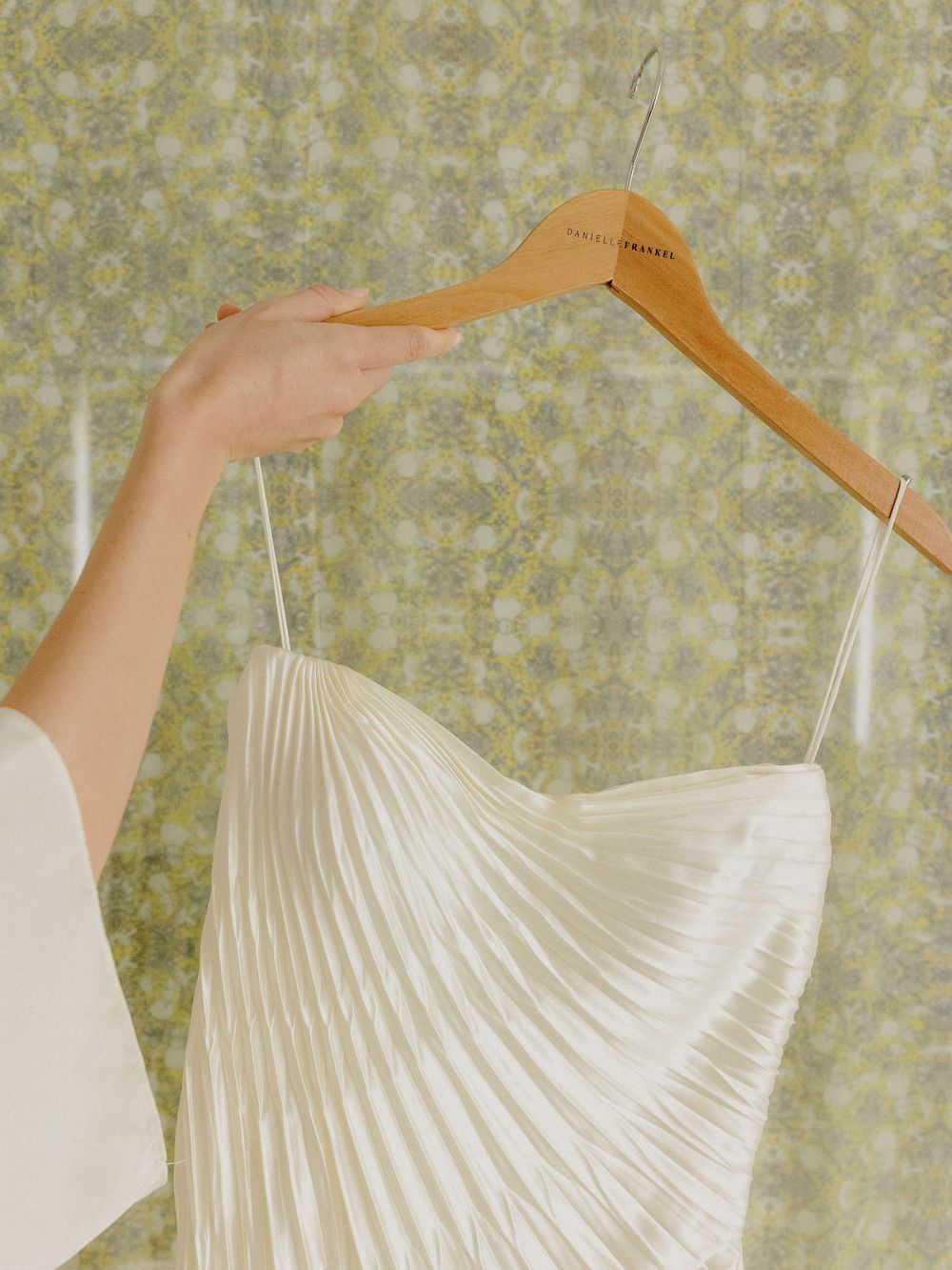 Close-up of a pleated white wedding dress bodice on a wooden hanger against floral wallpaper.