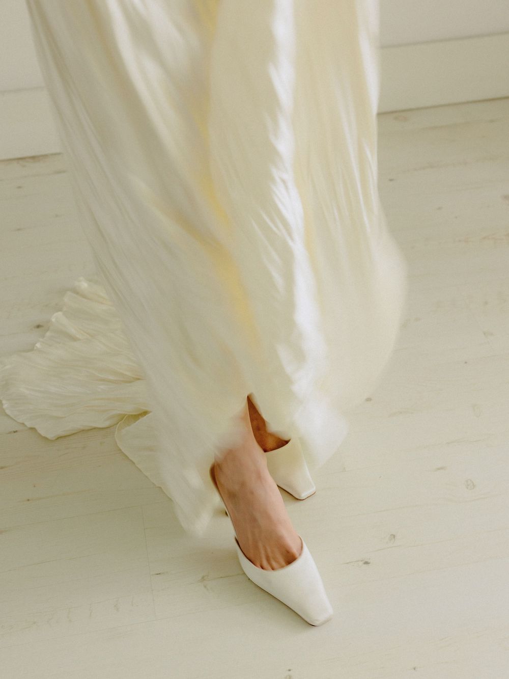 White bridal shoes peek out beneath flowing wedding dress hem on light wooden floor.