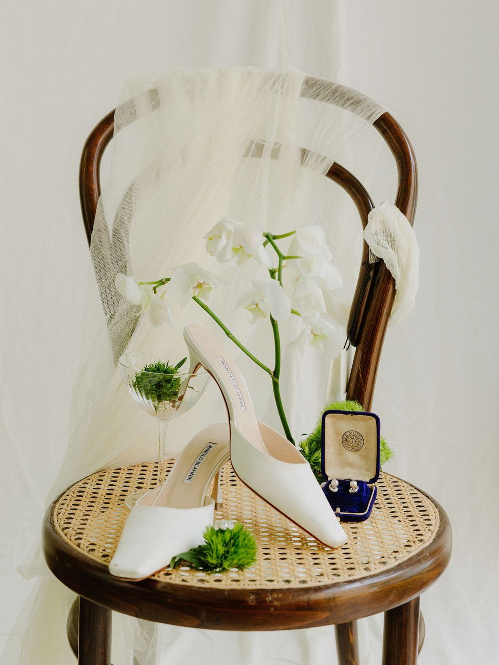 White bridal shoes and jewelry box displayed on a vintage cane chair with delicate floral accents.