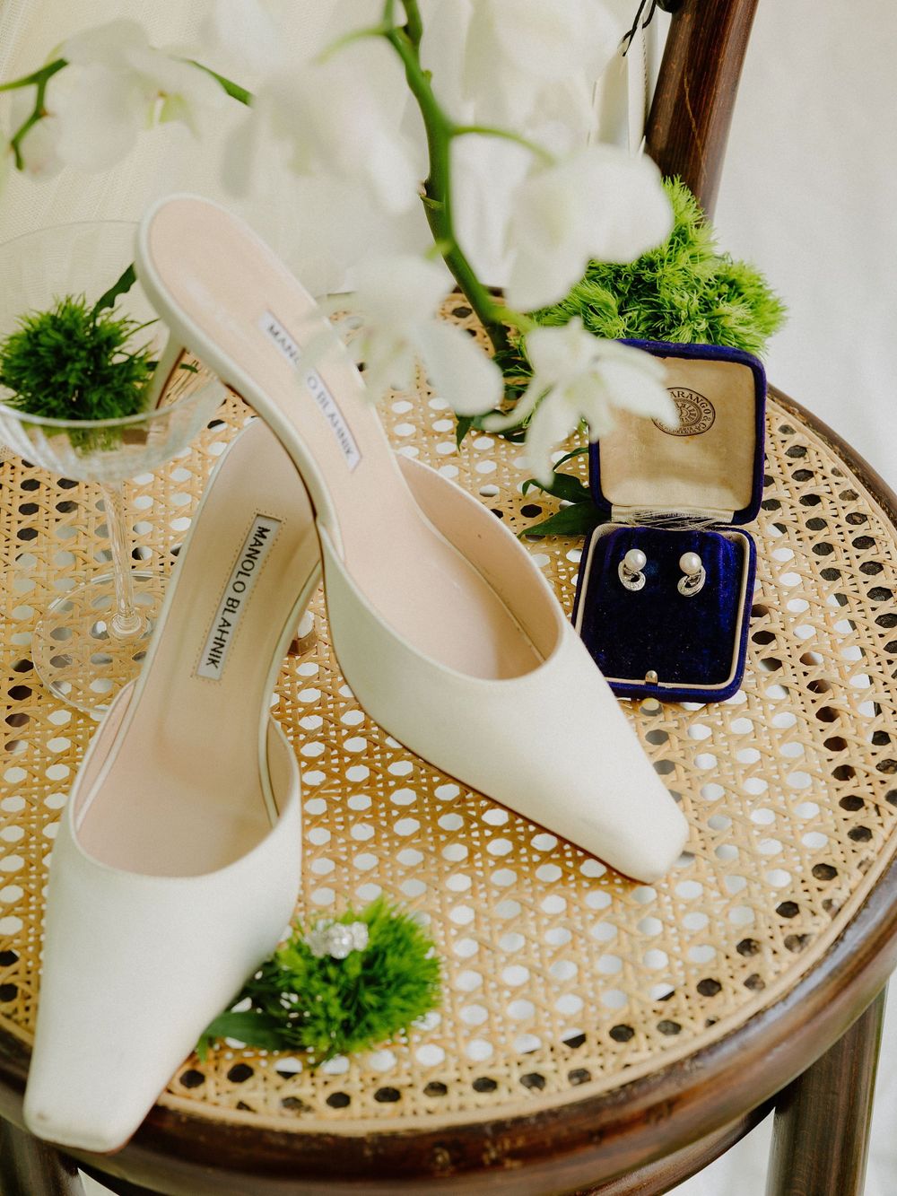 Elegant white wedding shoes and jewelry box arranged on a rattan chair with green floral decorations.