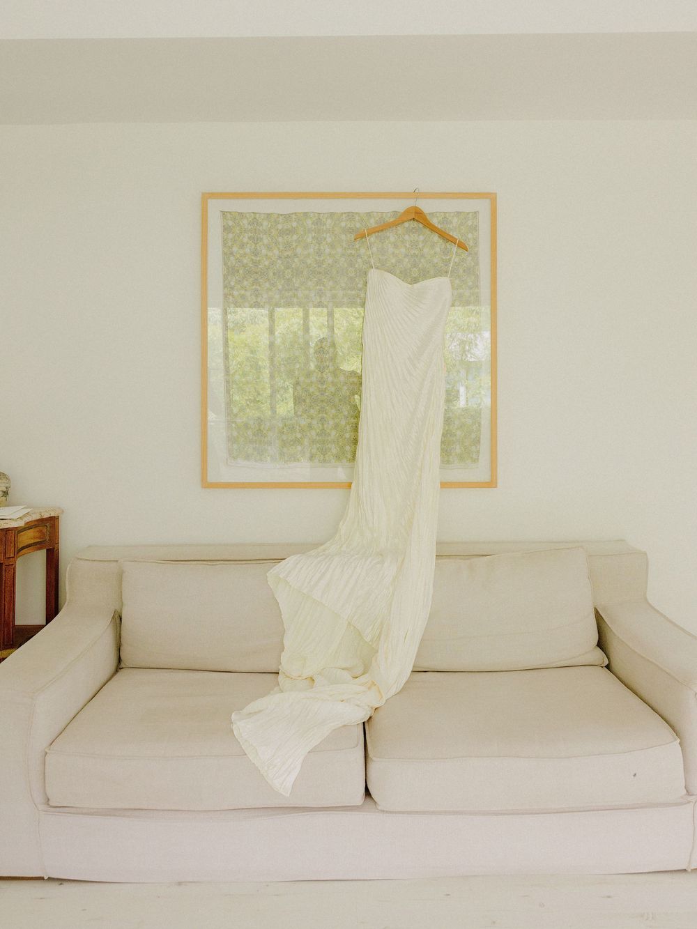 A white wedding dress hanging on display against a cream sofa and framed artwork.