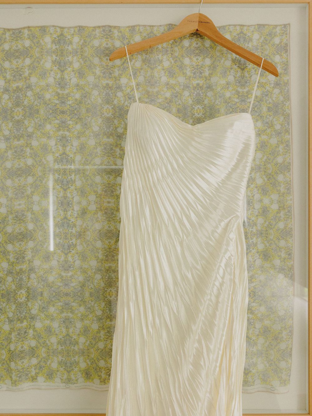 A pleated white wedding dress with sweetheart neckline hanging against patterned wallpaper.