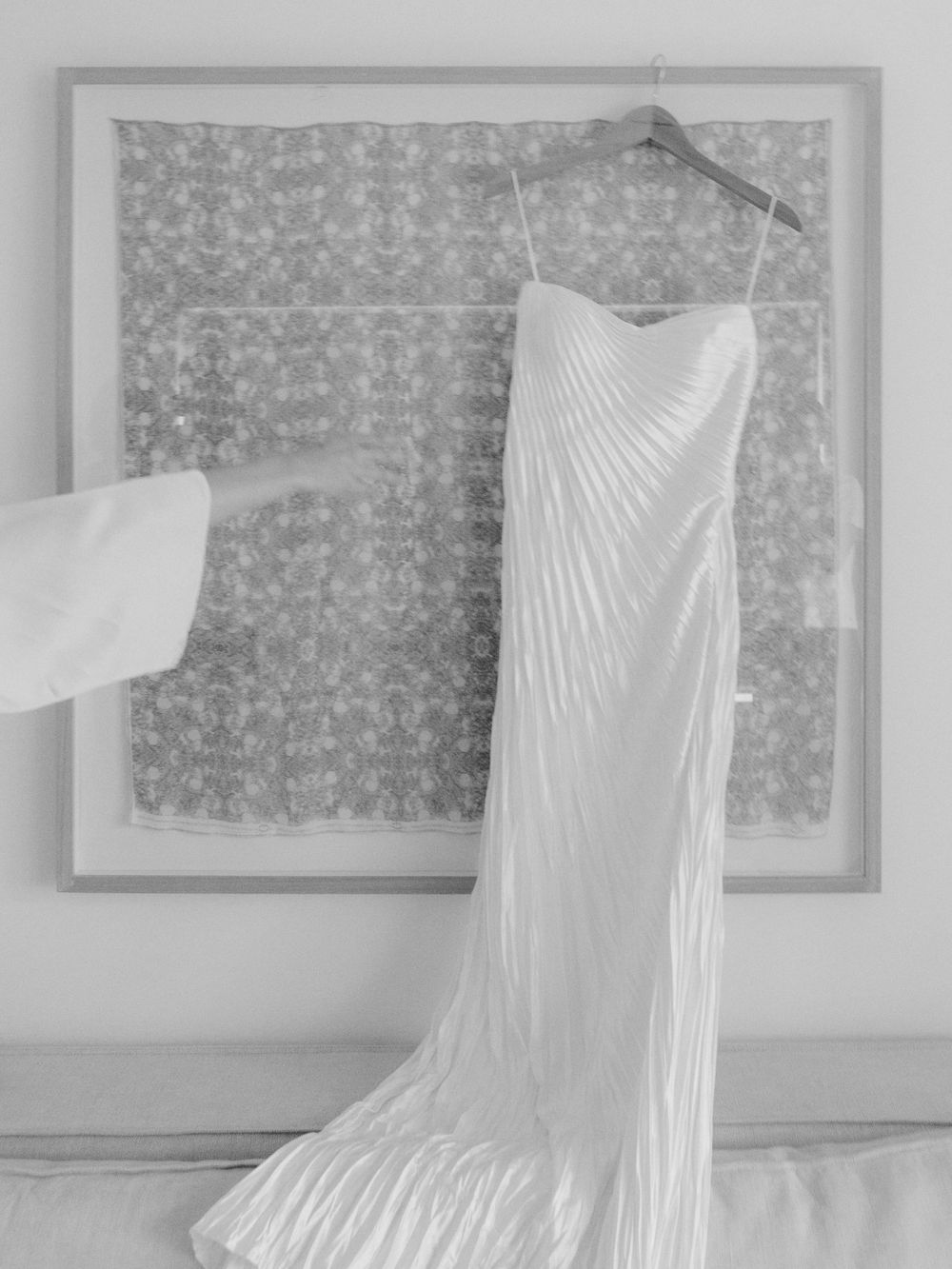 A minimalist white wedding gown with diagonal pleating displayed against a gray patterned background.