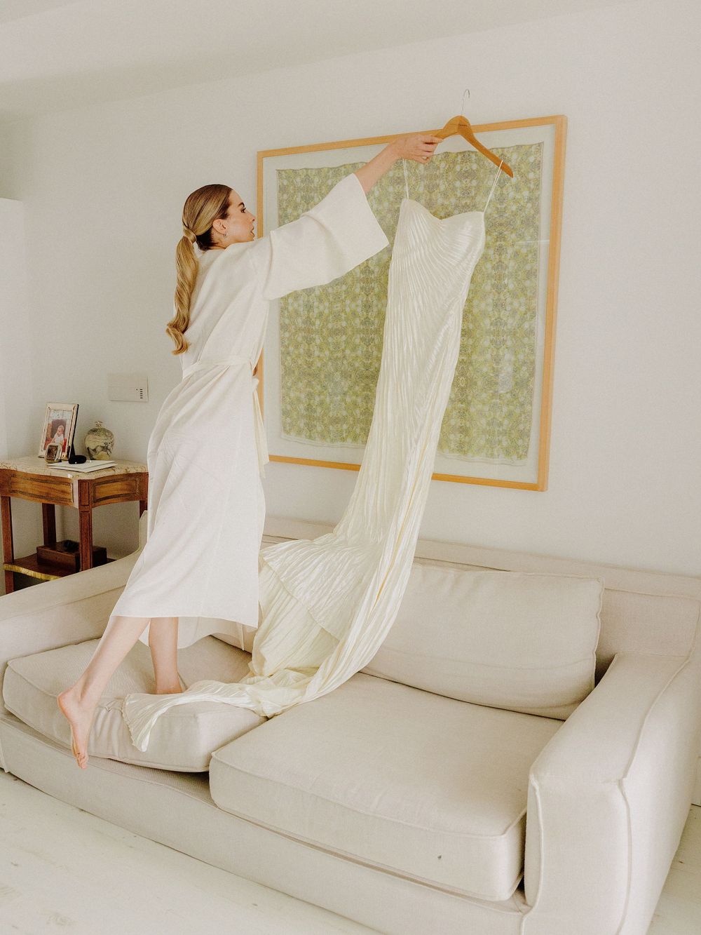 A person in white standing on a couch reaches up to adjust artwork on the wall in a minimalist setting.