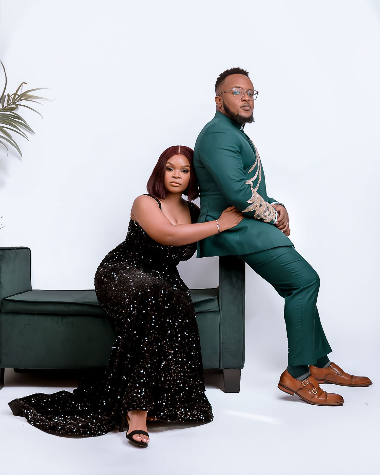 A couple poses in a studio setting with a black sparkly dress and green suit against a white backdrop with modern furniture.