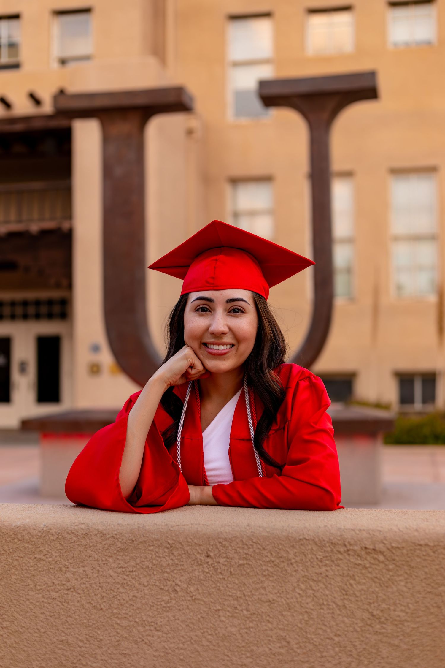 Graduate in red cap and gown poses at university building during sunset.