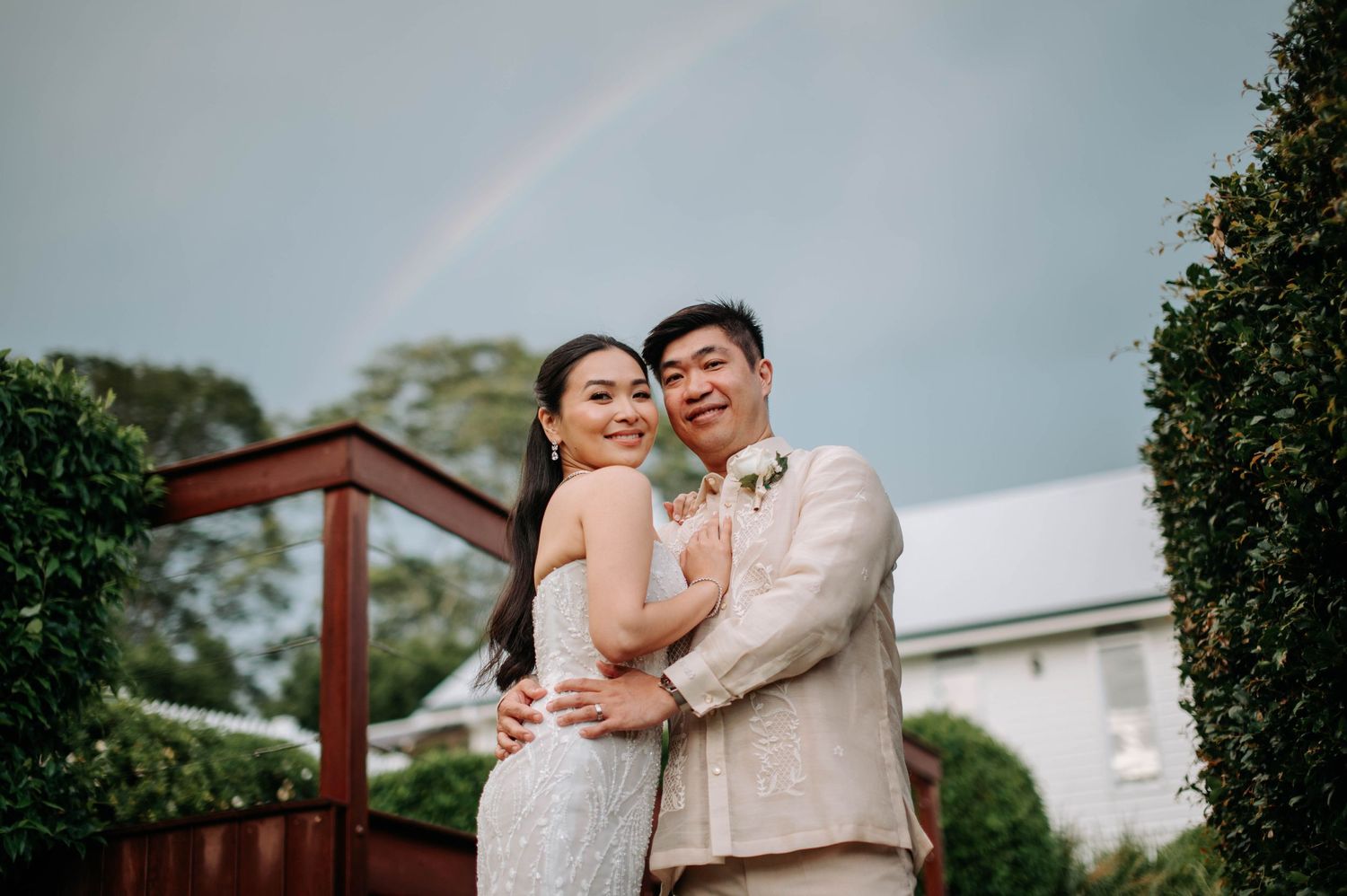 Couple embracing outdoors with rainbow and white building in background.