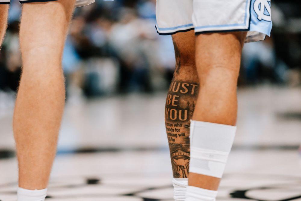 UNC basketball player with Just Be You tattoo on leg wearing light blue uniform and ankle wrap.