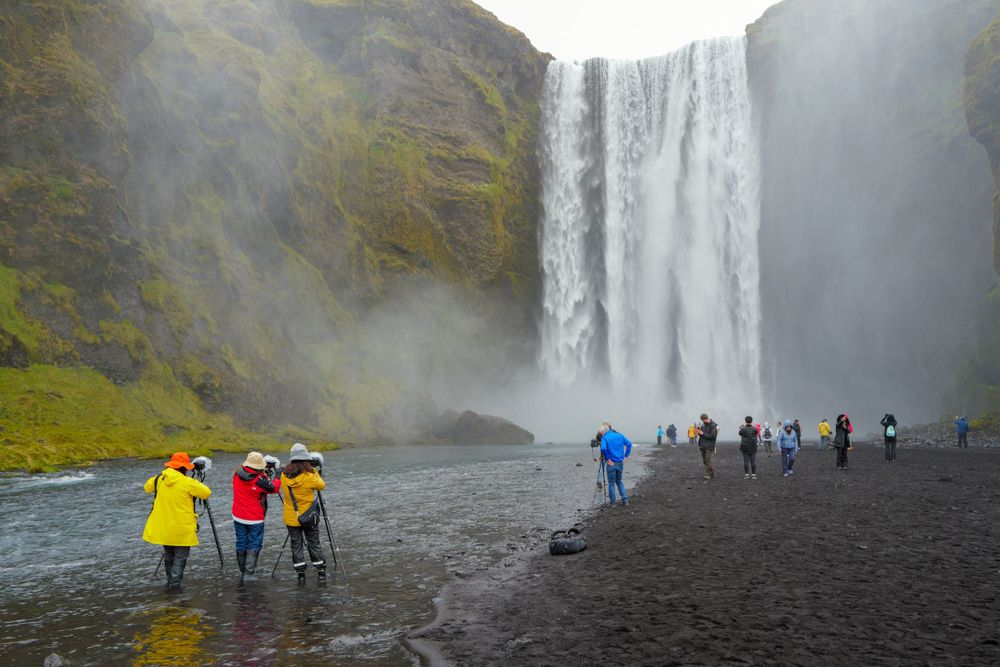 Tourists in colorful rain gear walk along black sand beach near massive Skogafoss waterfall in Iceland.