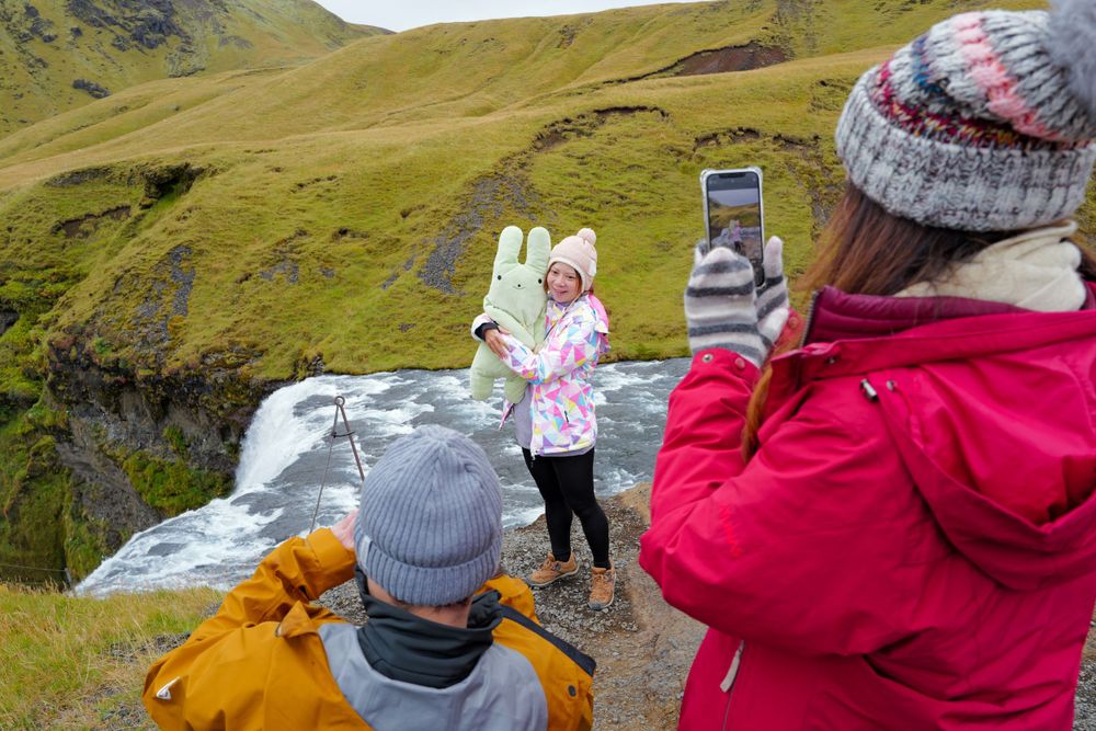 Tourists in winter jackets take photos of a scenic waterfall along green mountainous terrain in Iceland.