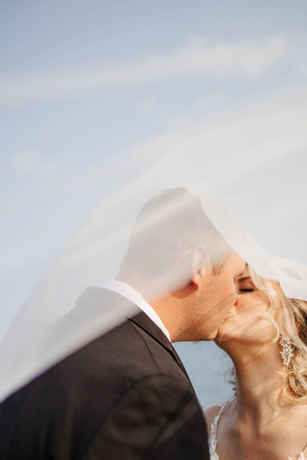 Newfoundland wedding photographer captures intimate close-up of couple kissing under flowing white veil against blue sky.