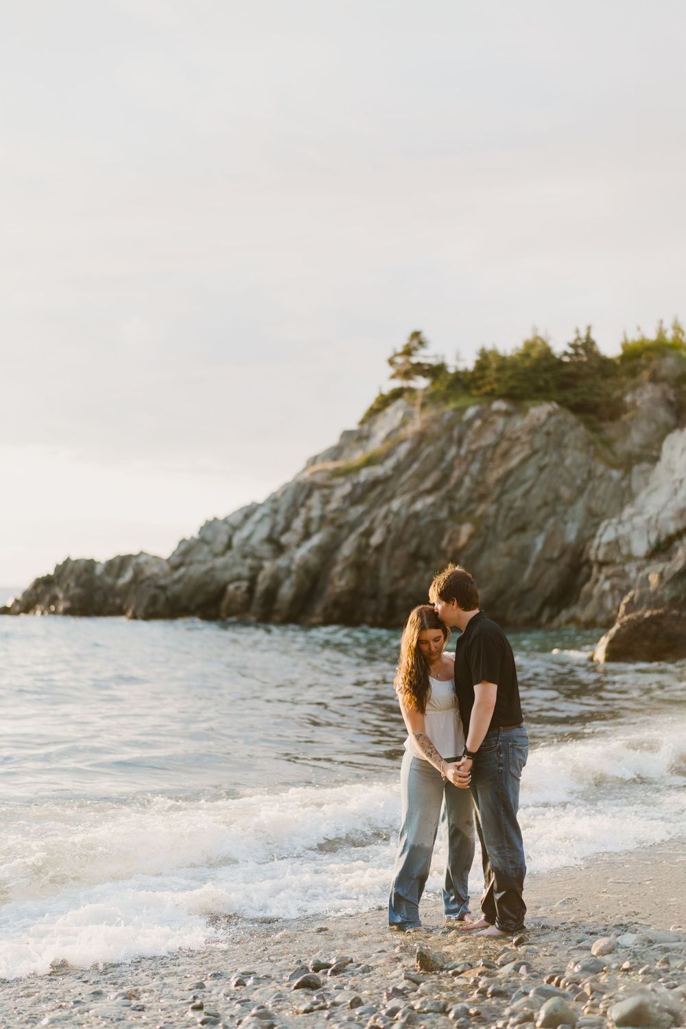 Newfoundland photographer captures romantic couple embracing on rocky beach at sunset with dramatic coastal cliffs.