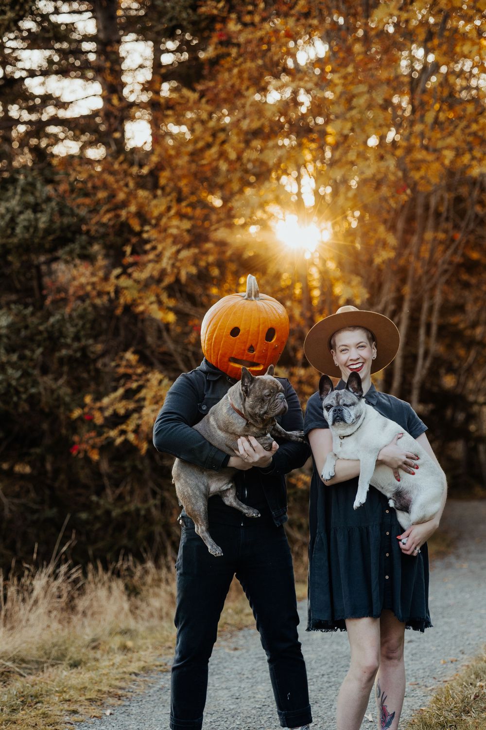 Couple in Halloween costumes holding dogs with jack-o-lantern head at sunset.