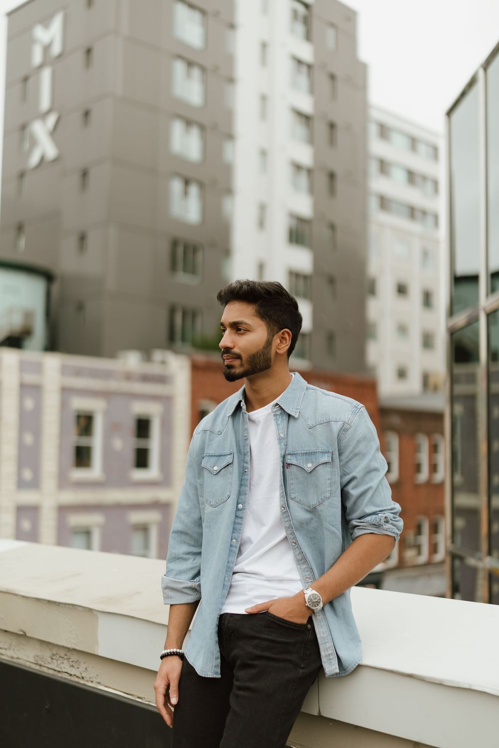 St. John's photographer captures casual man in denim shirt leans against urban rooftop wall with city buildings in background.