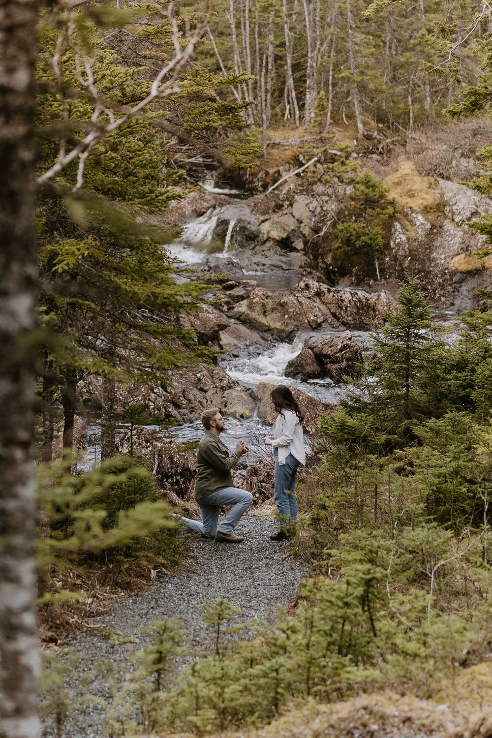 A scenic waterfall cascades down rocky terrain as people enjoy viewing it from a gravel path surrounded by evergreen trees.