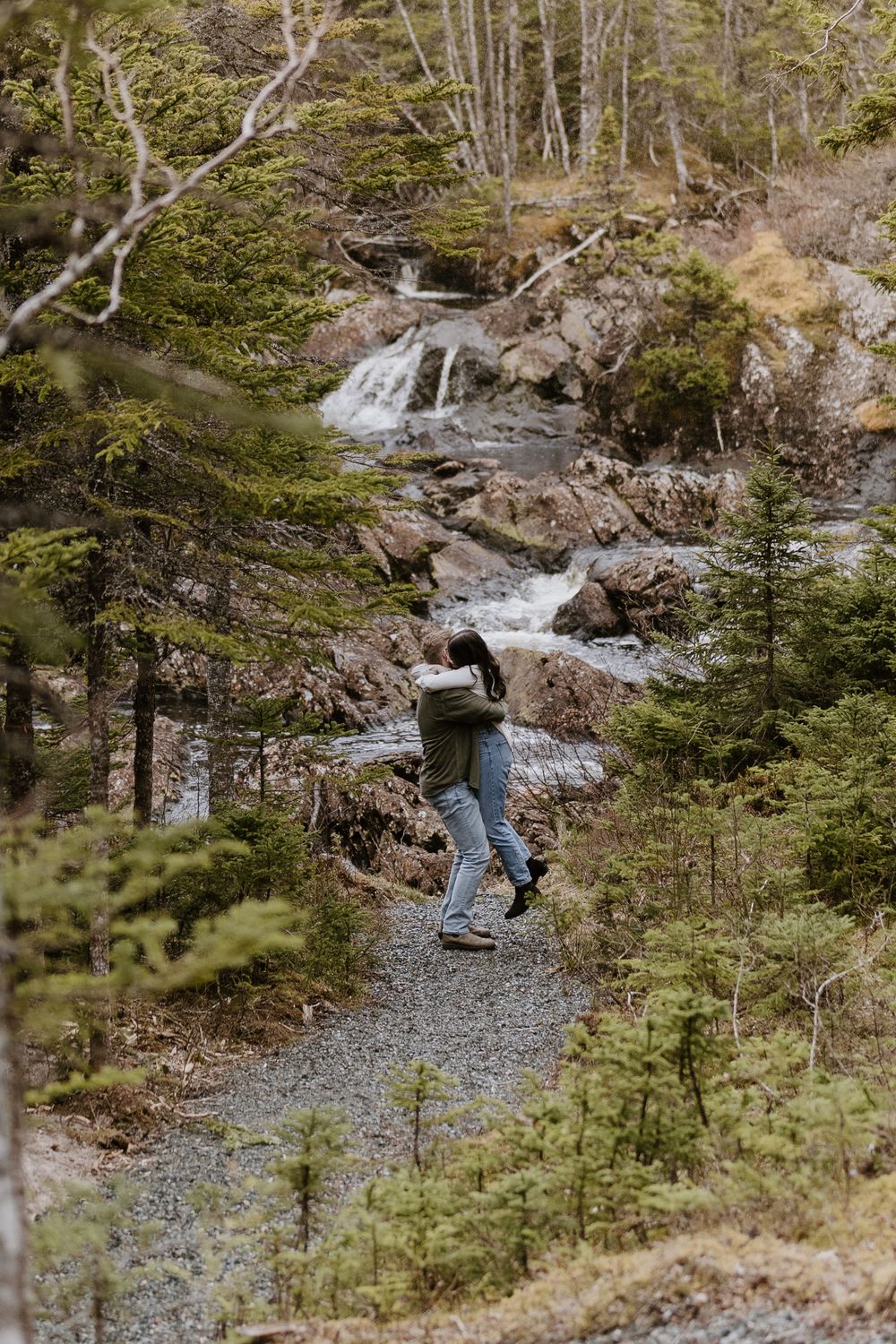 Cascading mountain stream flows through rocky forest terrain with evergreen trees and gravel paths.