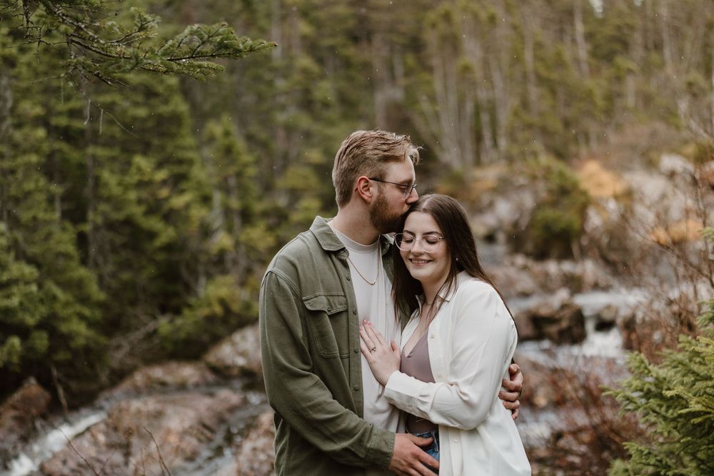 A couple shares an intimate moment outdoors against a scenic forest and stream backdrop in the wilderness.
