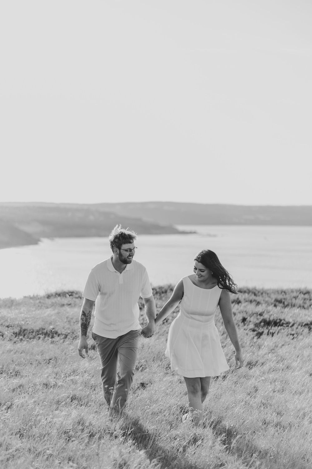 A couple runs playfully through a grassy meadow overlooking the ocean in a series of black and white photographs.
