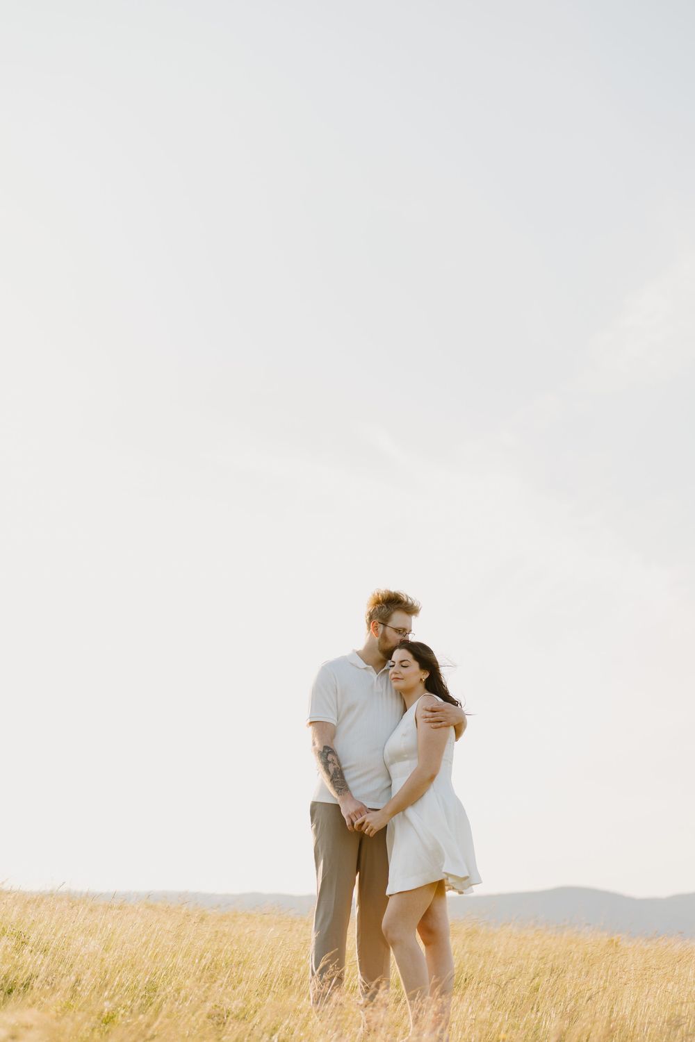 A couple embraces and kisses in a sunlit golden wheat field with mountains visible in the background.