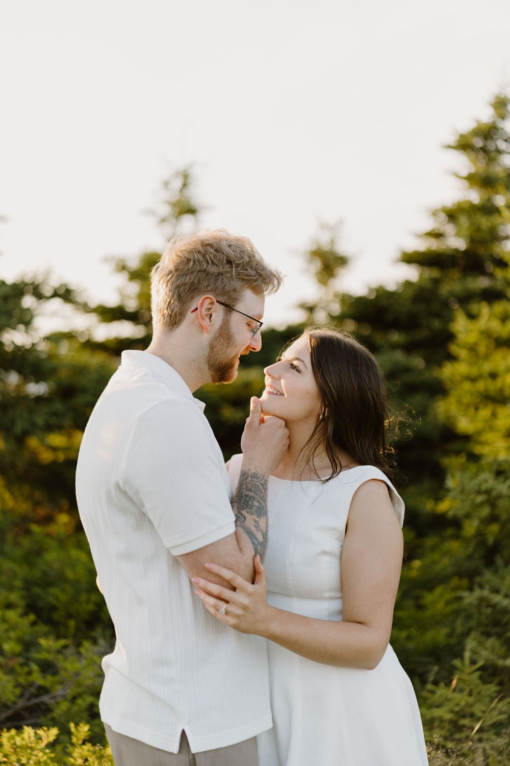 Happy couple dressed in white laughs together during golden hour photoshoot in a forest setting.