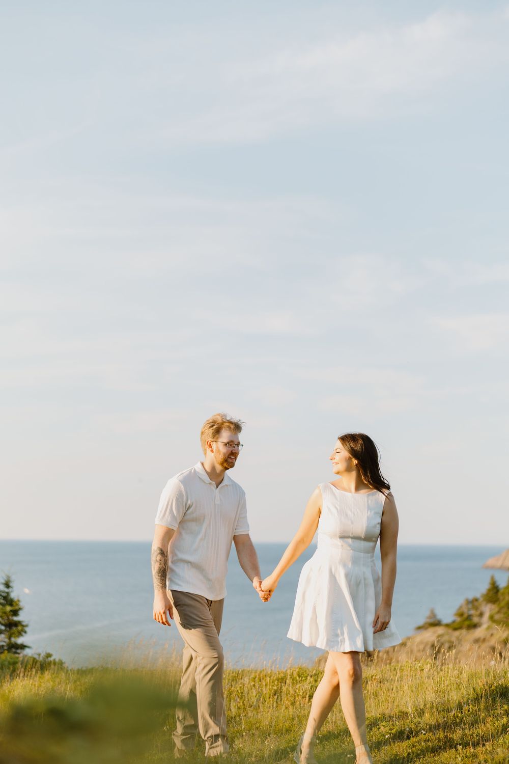 Couple in white outfits holding hands while walking through a scenic hilltop meadow at golden hour with ocean views.