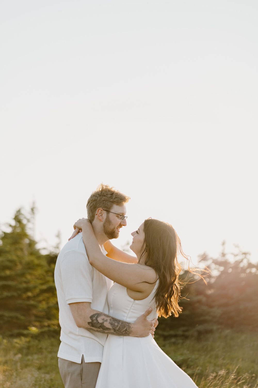 A romantic couple embraces during golden hour sunset with evergreen trees in the background.