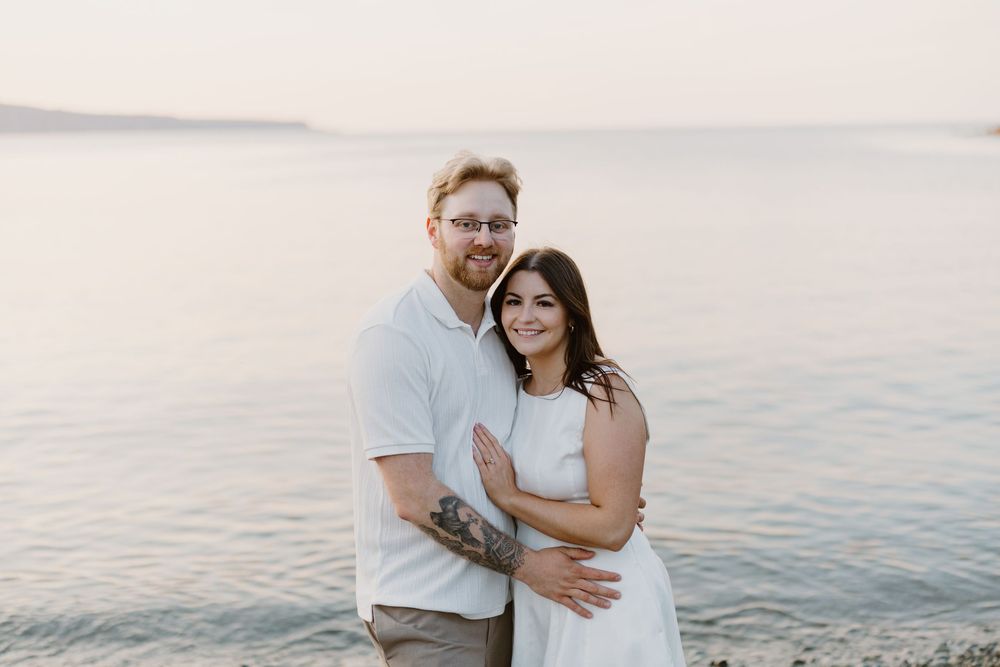A happy couple embraces while standing on a beach at sunset with calm water in the background.