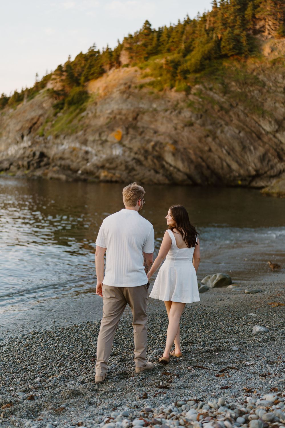 Couple walking hand in hand along a rocky beach with evergreen trees and cliffs visible in the background.