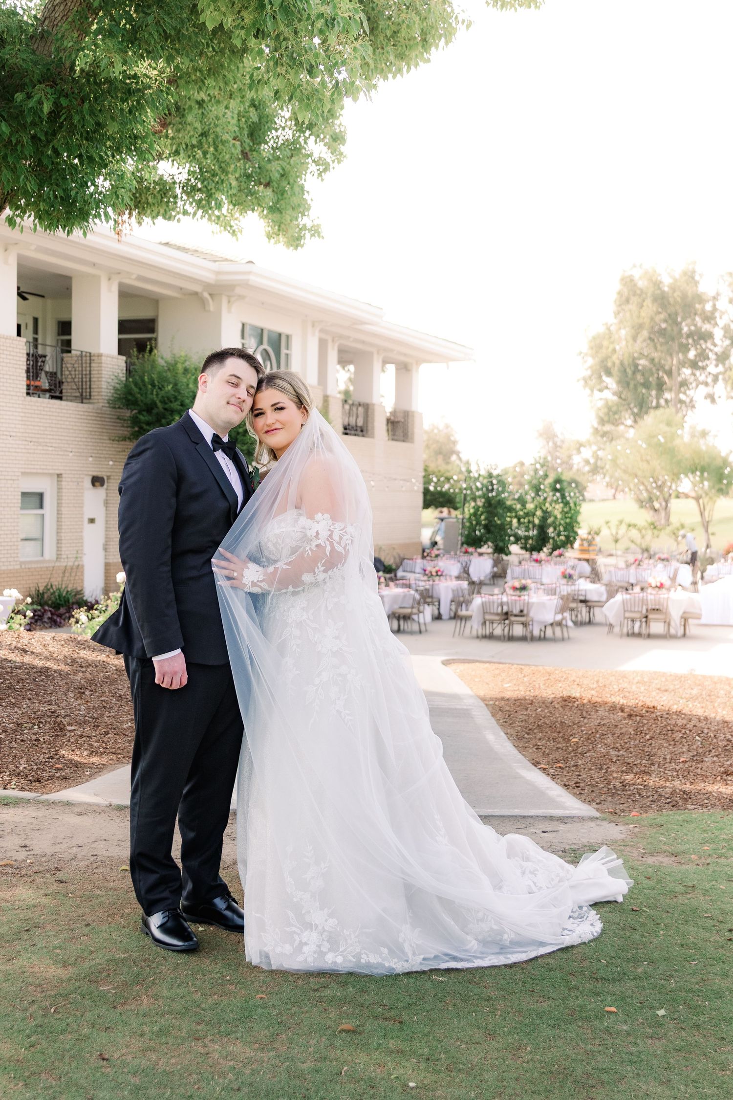 A couple poses for wedding photos in front of a grand white mansion with outdoor reception setup.