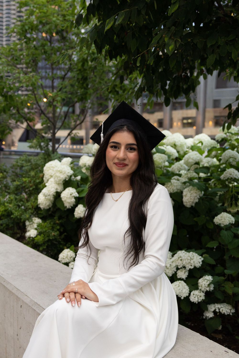 Young woman in white dress and graduation cap poses outdoors near white hydrangeas with city buildings in background.