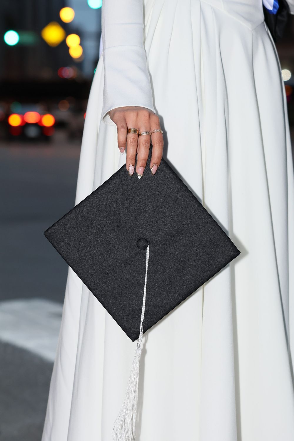 Graduate in white gown holding black mortarboard cap with white tassel on city street at night.