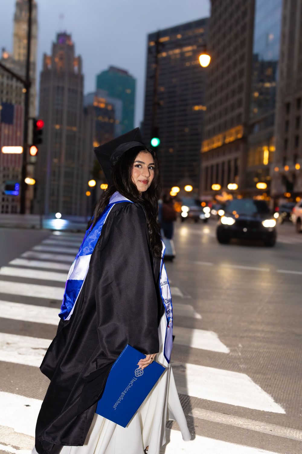 Graduate in black cap and gown with blue sash crosses city street at dusk, holding diploma in urban downtown setting.