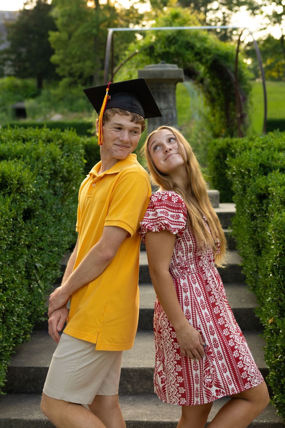 A smiling young man in a graduation cap and yellow polo poses back-to-back with a blonde girl in a red dress outdoors.
